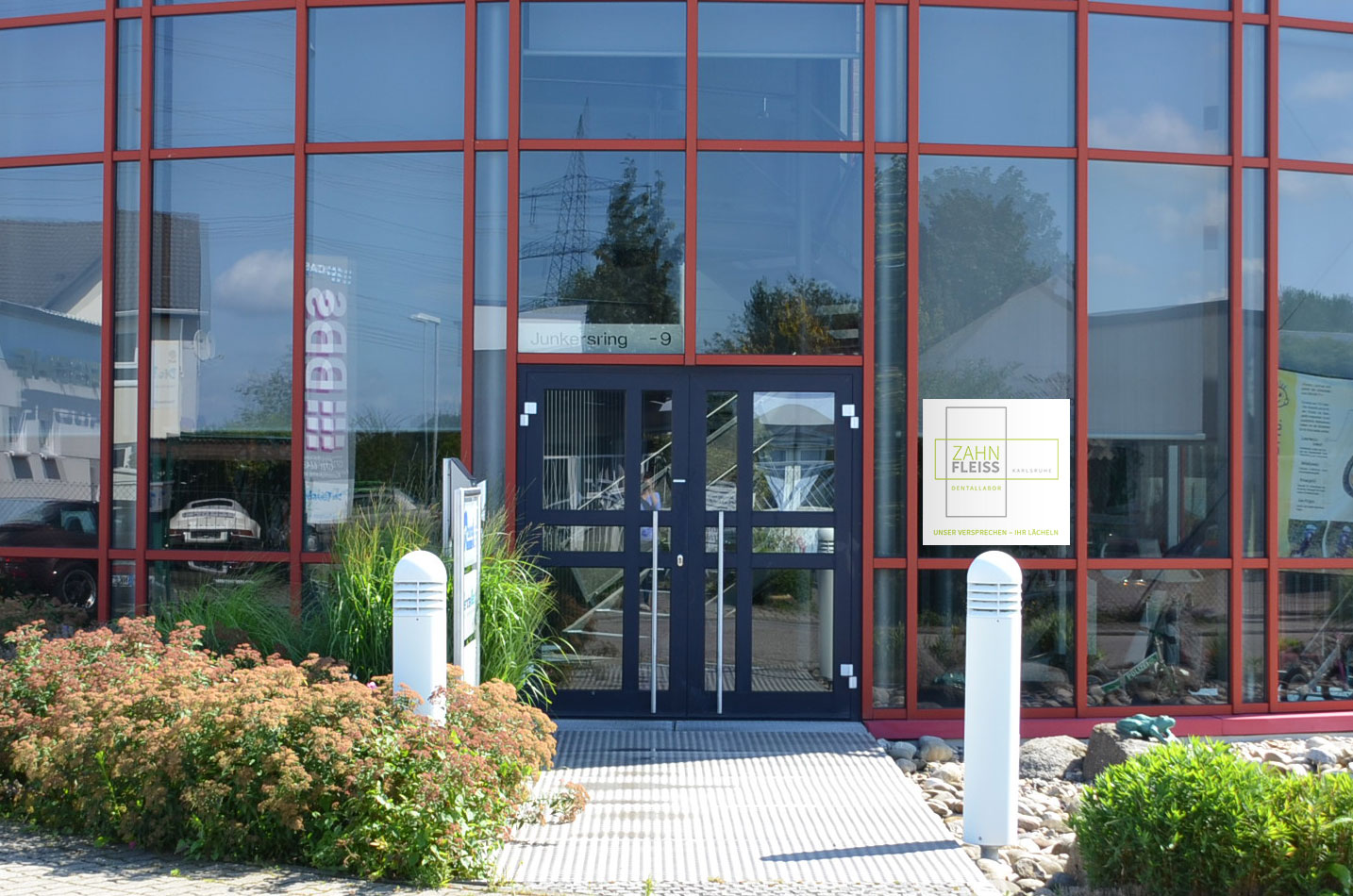 Ein modernes Bürogebäude mit großen Fenstern, roten Rahmen und einer Eingangstür, umgeben von gepflegten Grünflächen.